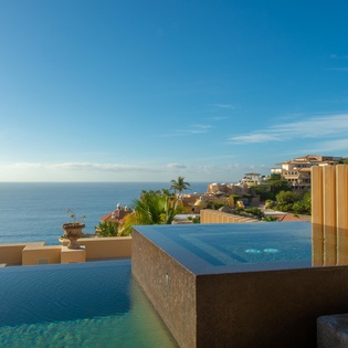 Infinity pool with stunning ocean views overlooking coastal villas and tropical palms. Relax poolside surrounded by serene blue skies and hillside scenery perfect for soaking in the coastal beauty.
