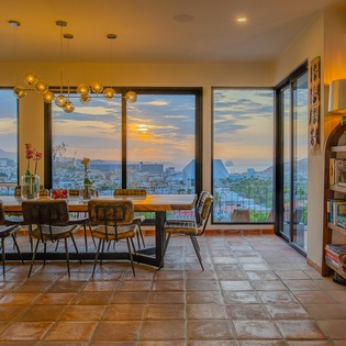 Gather around this inviting dining table as warm amber lights glow overhead and watch the sun paint the sky in golden hues through floor-to-ceiling windows overlooking the cityscape.