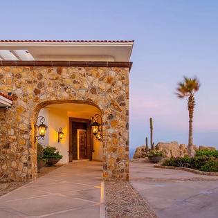 Stone archway entrance with glowing lanterns welcomes arrivals at this coastal villa as dusk settles over palm trees and ocean views beyond.