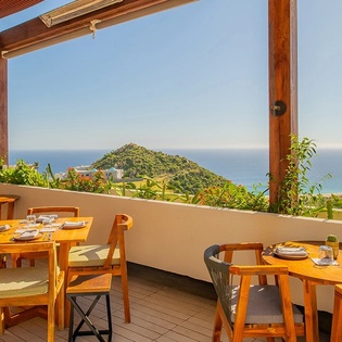 Outdoor dining terrace with panoramic sea and mountain views, featuring wooden furniture under a pergola.