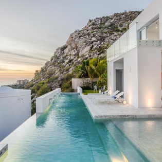 Modern hillside villa with infinity pool overlooking dramatic rocky cliffs and distant coastline at golden hour.