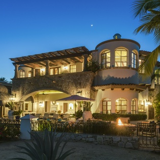 Stunning Mediterranean-style villa illuminated under twilight skies, surrounded by tropical palms and mature landscaping in an upscale beachside setting.