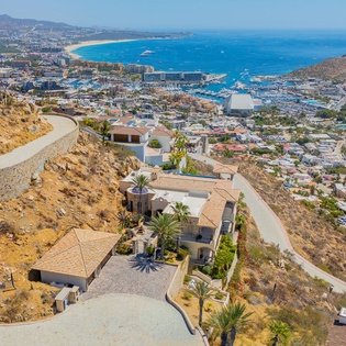 Stunning aerial view of hillside villa overlooking coastal town and turquoise sea with pristine beach and mountain backdrop.