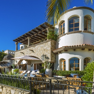 Stunning Mediterranean villa with stone accents and curved architecture, surrounded by tropical palms and lush landscaping under brilliant blue skies.