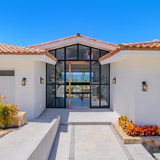 Modern luxury villa featuring striking glass entrance, terracotta tile roof, and landscaped walkway under brilliant blue skies.
