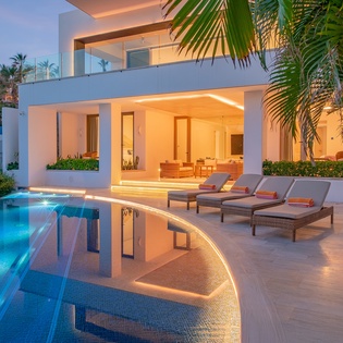Modern tropical villa featuring a curved swimming pool, elegant lounge chairs, and lush palm trees in a serene beachside setting.