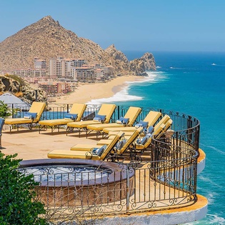Elevated terrace featuring sun loungers and coastal panorama with dramatic rocky headland, pristine beach, and turquoise waters dotted with boats in the distance.