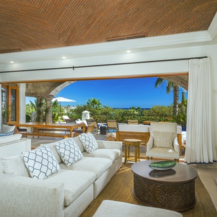 Relax in your elegant living room where soaring brick ceilings and flowing white curtains frame stunning tropical views through panoramic doors.