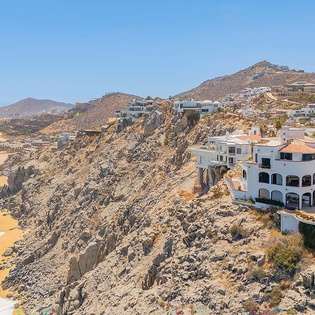 A dramatic cliffside property overlooks golden beaches and the Pacific coastline, nestled among rocky hillsides and palm trees in this coastal community.
