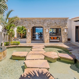 A stone walkway leads you across a serene pool toward a stunning stone-and-stucco pavilion framed by swaying palms and ocean views. Your private coastal oasis awaits.