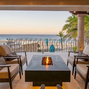 Unwind on your private terrace with plush seating surrounding a warm fireplace, where palm trees frame stunning ocean views and gentle evening light creates the perfect ambiance for relaxation.