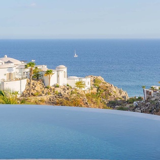 Coastal property with infinity pool overlooking blue ocean waters, surrounded by white Mediterranean-style architecture, palm trees, and rocky hillside terrain—a scenic seaside setting with a sailboat in the distance.