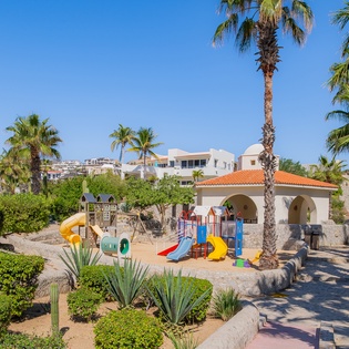 Colorful playground with slides and climbing equipment surrounded by palm trees and landscaped gardens in a sunny Mediterranean setting.