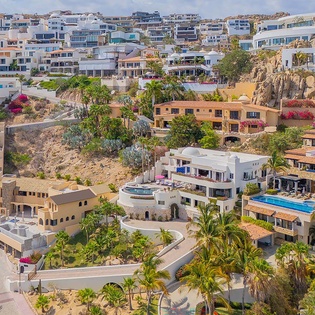 An aerial view showcasing the property's hillside location among lush palm trees and neighboring vacation homes, set against a rocky coastal landscape.