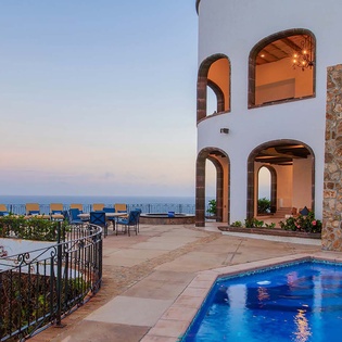 A coastal villa with Mediterranean architecture features arched windows, stone accents, and a private pool overlooking the sea at twilight.