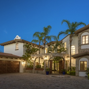 Elegant Mediterranean-style villa with stone accents and palm trees, illuminated against the evening sky.