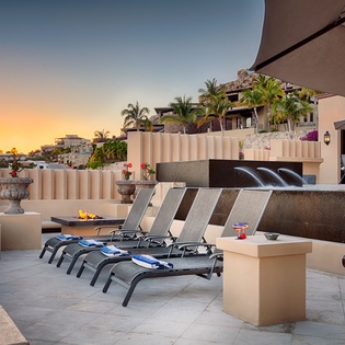 A rooftop terrace overlooking a hillside resort community at golden hour, featuring lounge chairs, a fire feature, and contemporary water elements with tropical palms in the distance.