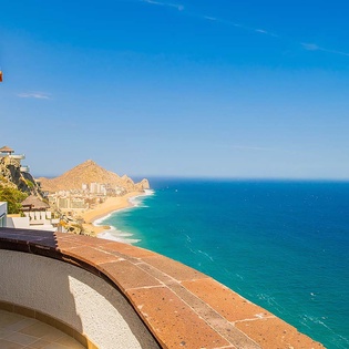 Coastal property overlooking turquoise ocean waters, sandy beach, and dramatic desert landscape under clear blue skies. Terracotta tile rooflines and hillside architecture frame this stunning Mexican Riviera setting.