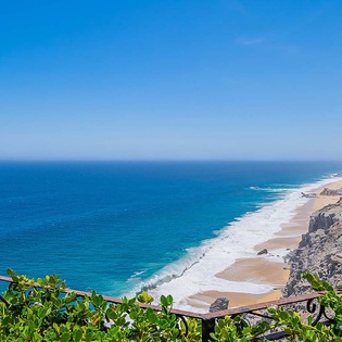The property overlooks a dramatic coastline where golden sands meet turquoise waters, framed by rugged cliffs stretching along the horizon.