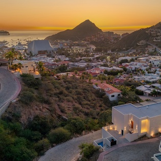 Stunning coastal hillside community features modern white villas overlooking azure waters during golden hour, with dramatic mountain backdrop and cruise ship views.
