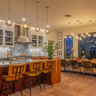 Gather around the warm wood island or dining table and soak in stunning city views through floor-to-ceiling windows. Your evenings here blend modern style with comfort, lit by ambient pendant lights and framed by a twilight skyline.