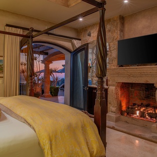 Unwind in your elegant four-poster bedroom where crackling fireplace warmth meets luxurious comfort, with stunning outdoor views through grand archway doors.