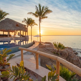 Beachfront property with infinity pool, thatched palapa, and palm trees overlooking the ocean at sunset, offering direct waterfront access and tropical coastal scenery.