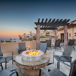 An oceanfront terrace overlooking Cabo's coastline features a stone fire pit surrounded by cushioned seating, perfect for sunset gatherings. The outdoor living space flows seamlessly to indoor comfort beneath a stylish pergola.