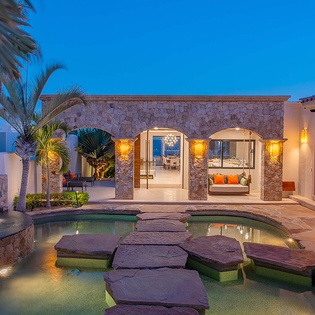 This luxurious property features a stunning stone-built courtyard illuminated by warm ambient lighting at dusk, with elegant archways opening to the main living areas and a serene stepping-stone water feature surrounded by tropical palms.