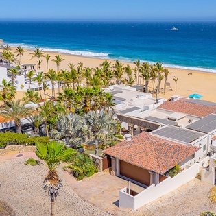 Aerial view of beachfront property nestled among swaying palms, overlooking golden sand and turquoise waters. A stunning coastal setting with dramatic rocky cliffs and endless ocean horizons.