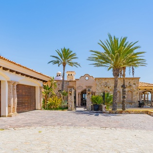 This stunning Mediterranean villa showcases dramatic stone architecture with panoramic ocean views, surrounded by swaying palms under endless blue skies.