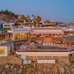 Luxury hillside villa perched on dramatic rocky coastline, featuring multiple levels with pools, terraces and thatched-roof palapas overlooking the sea.