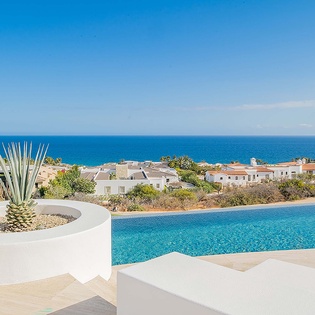 The infinity pool stretches toward the brilliant blue ocean, framed by sun-bleached architecture and desert plants—your perfect spot to soak up the Mediterranean sunshine.