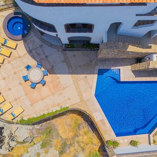Aerial view of the property showcasing two vibrant pools, a circular spa, and a curved sunbed terrace bordered by natural coastal landscape.