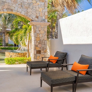 Your private terrace invites you to sink into modern lounge chairs with vibrant orange cushions, framed by palm trees and a stone archway leading to the garden and pool beyond.