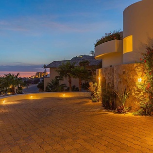 The property's illuminated facade welcomes arrivals at twilight, with Mediterranean-style architecture and ocean views visible from the surrounding hillside location.