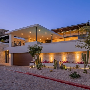 Modern luxury villa with dramatic lighting showcases contemporary architecture surrounded by desert landscaping and mountain views.