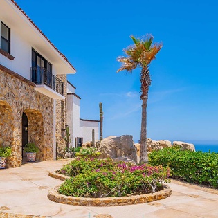 Stunning stone villa entrance with ocean views, framed by tropical palms and colorful flowering gardens under brilliant blue skies. A welcoming coastal retreat with authentic architectural charm.