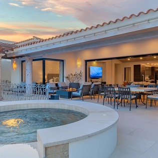 An elegant outdoor terrace featuring a hot tub, dining area, and lounge seating that welcomes you with warm lighting and stunning sunset views over the landscape.