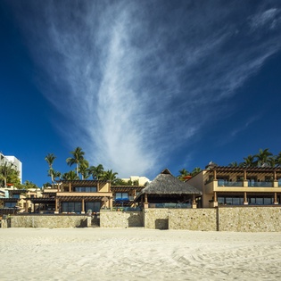 Beachfront resort properties with traditional architecture line the pristine sandy shore under dramatic skies.