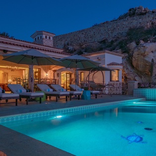 Luxury villa with illuminated swimming pool set against dramatic desert mountains, featuring elegant Mediterranean architecture and premium outdoor lounging areas.