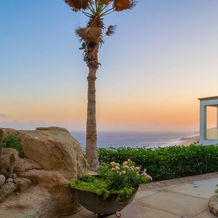 The property features a coastal desert landscape with native cacti, palm trees, and colorful garden plantings, offering stunning views of the ocean and surrounding area at sunset.