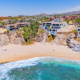 Stunning aerial view of beachfront properties nestled between golden sand and turquoise waters, with mountains providing a dramatic backdrop.