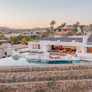 An elevated view of a stunning villa showcasing its contemporary design, terraced landscaping with natural stone walls, and luxurious outdoor living spaces set against mountains and tropical palms.