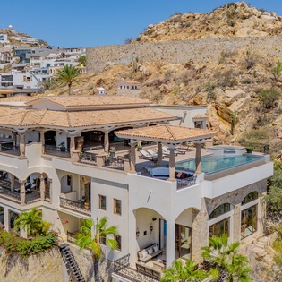 Stunning hillside villa showcasing Mediterranean architecture with rooftop pool amid dramatic desert landscape and neighboring coastal community.