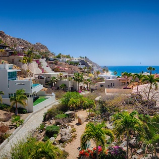 Coastal hillside community featuring terraced homes overlooking the ocean, with palm trees dotting the rocky landscape and a winding pathway through landscaped gardens. The property area showcases colorful architecture against dramatic blue skies.