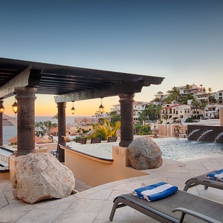 A stunning hillside resort pool area with stone columns and Mediterranean-style architecture captures the golden glow of sunset over coastal cliffs.