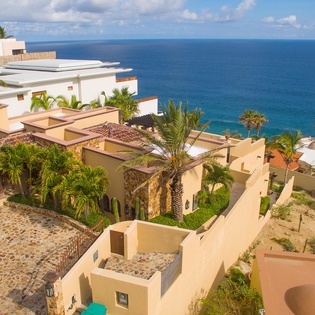 An aerial view showcases the property's beachfront location, with terracotta-roofed villas, lush tropical landscaping, and the stunning turquoise ocean beyond. The resort is nestled along the coastline in a serene, picturesque setting.