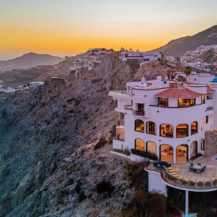 A stunning coastal estate perched on dramatic cliffs overlooking the ocean, captured during a breathtaking sunset. The property features hillside architecture, a private pool, and panoramic views of the beach and surrounding mountains.