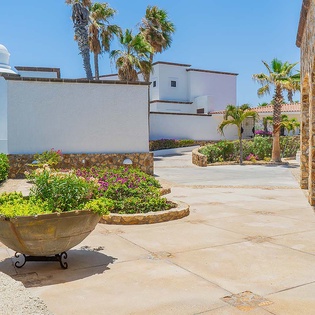 Sun-drenched courtyard with vibrant gardens, rustic stone columns, and palm trees leading to white-washed villa entrances—a welcoming arrival to a serene coastal retreat.